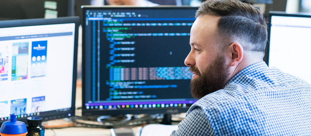 The image shows a software develop working at a desk, on which are multiple monitors of code.