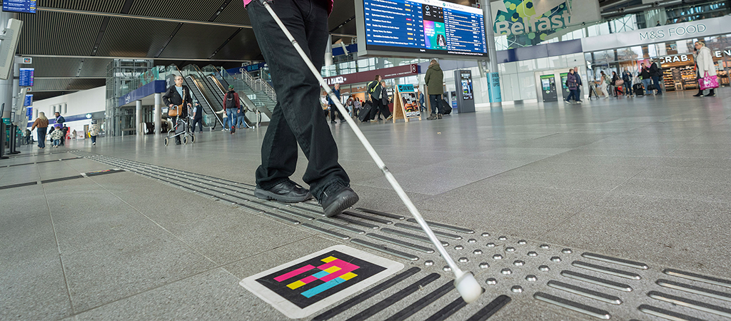 The image shows a user with a guide cane using tactile flooring and NaviLens codes to navigate Belfast Grand Central Station
